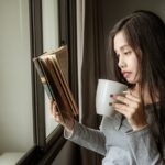 A woman holds an aged book and a white mug beside a bright window.